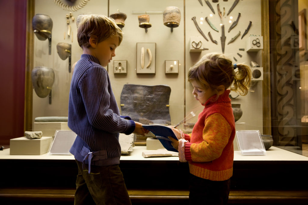 boy and little girl at excursion in historical museum near exhibits of ancient relics in glass cases, girl writes to writing-books